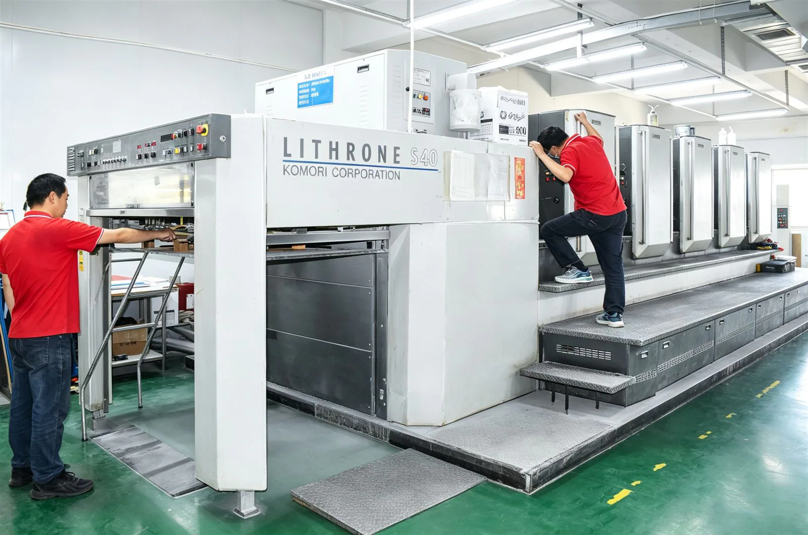 Two operators at the Lithrone press on the factory floor, holding color and registration on branded scratcher sheets.